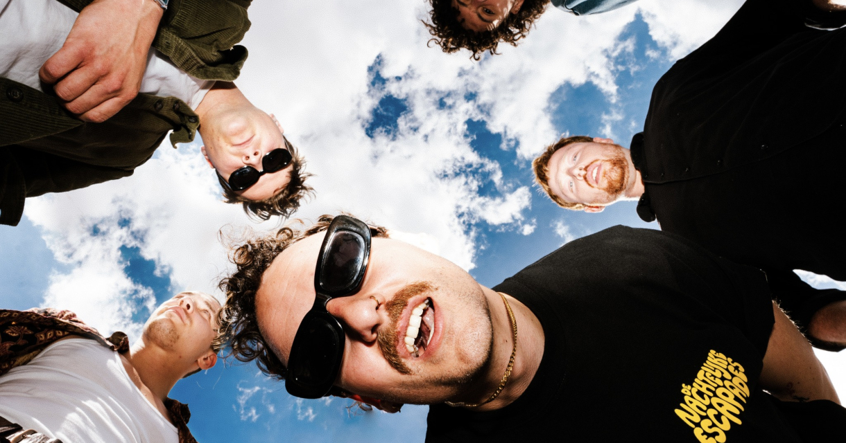 Vijf vrienden in een cirkel, kijkend naar de camera met een blauwe lucht op de achtergrond.