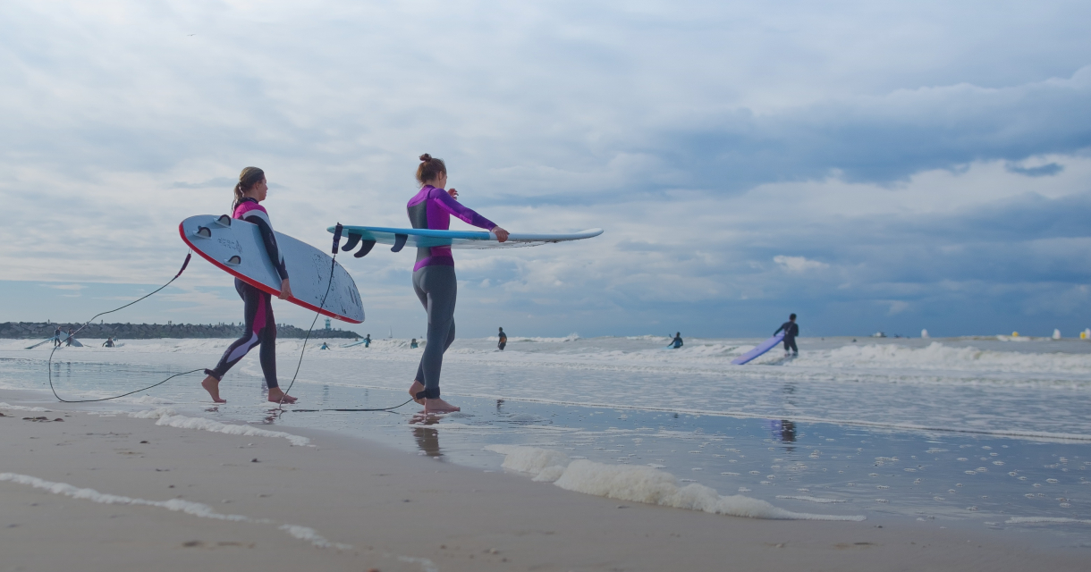 Twee surfers met surfplanken lopen langs het water.