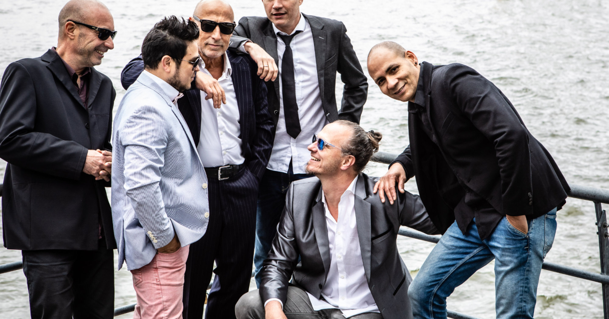 Zes mannen poseren aan de oever van een waterlichaam.