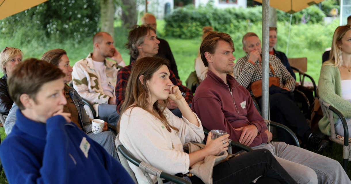 Groep mensen zit onder een parasol en luistert aandachtig.