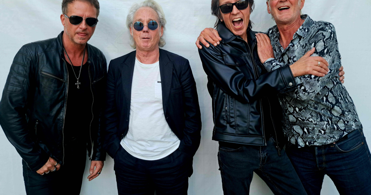 Vier mannen in casual outfits en zonnebrillen poseren vrolijk samen.