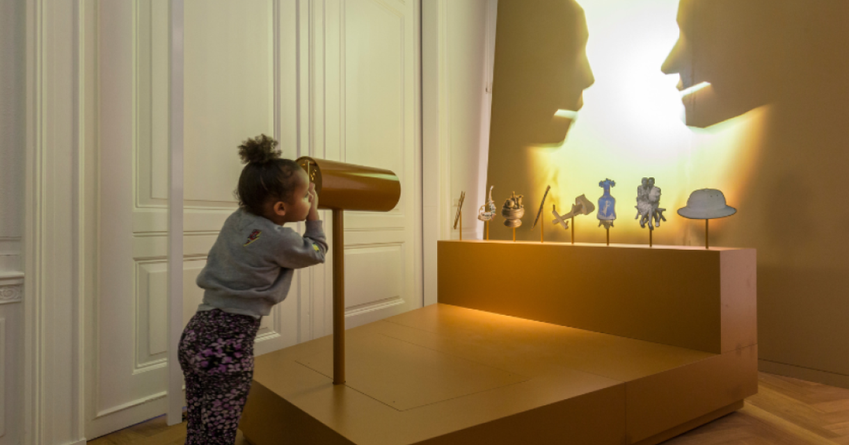 Een kind kijkt door een gouden kijkglas bij een expositie met silhouetten en voorwerpen.