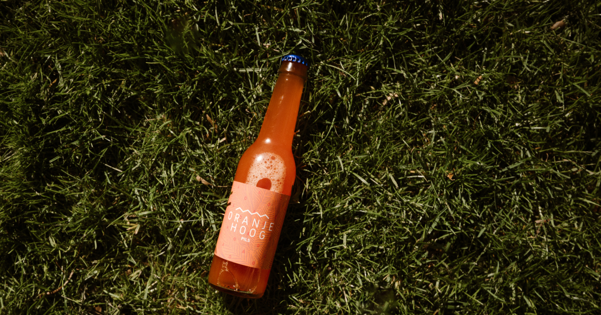 Een fles Oranje Hoog pils ligt op het gras.