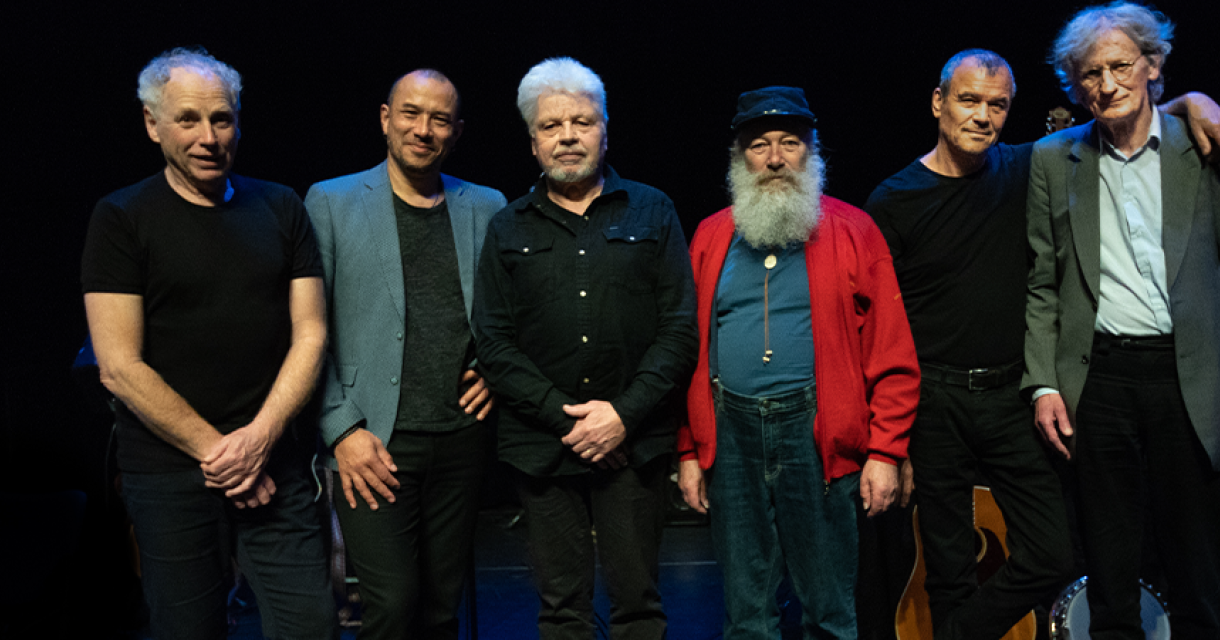 Zes muzikanten poseren samen op het podium.