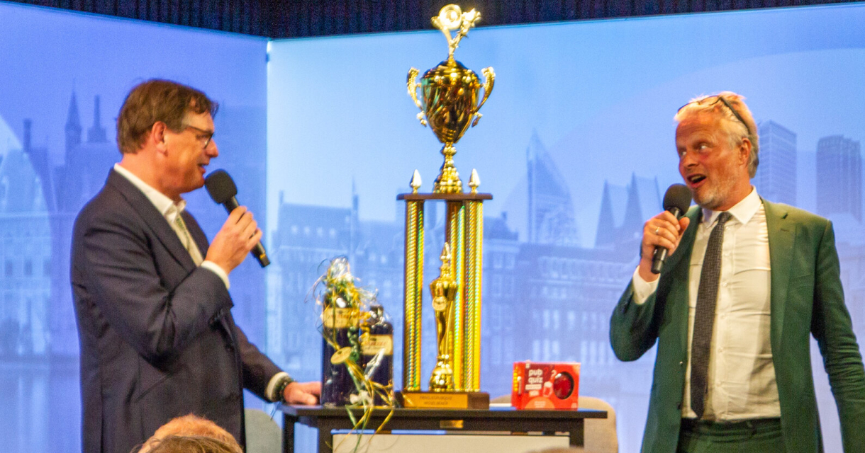 Twee mannen presenteren een trofee op een podium.