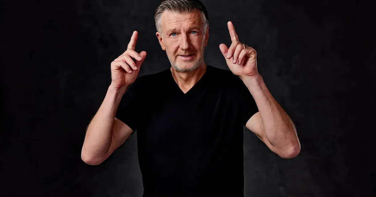 Een man in een zwart shirt wijst omhoog met beide handen.
