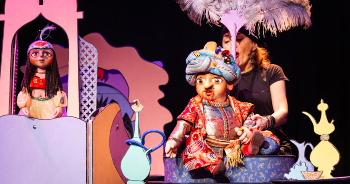 Twee kleurrijke marionetten op een podium, een met een kleurrijk kostuum en een hoed, de ander in traditionele kleding.