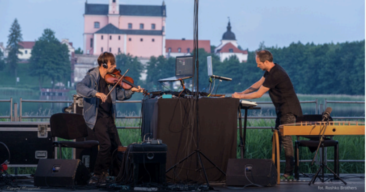 Twee muzikanten spelen op een podium bij een meer met een kasteel op de achtergrond.