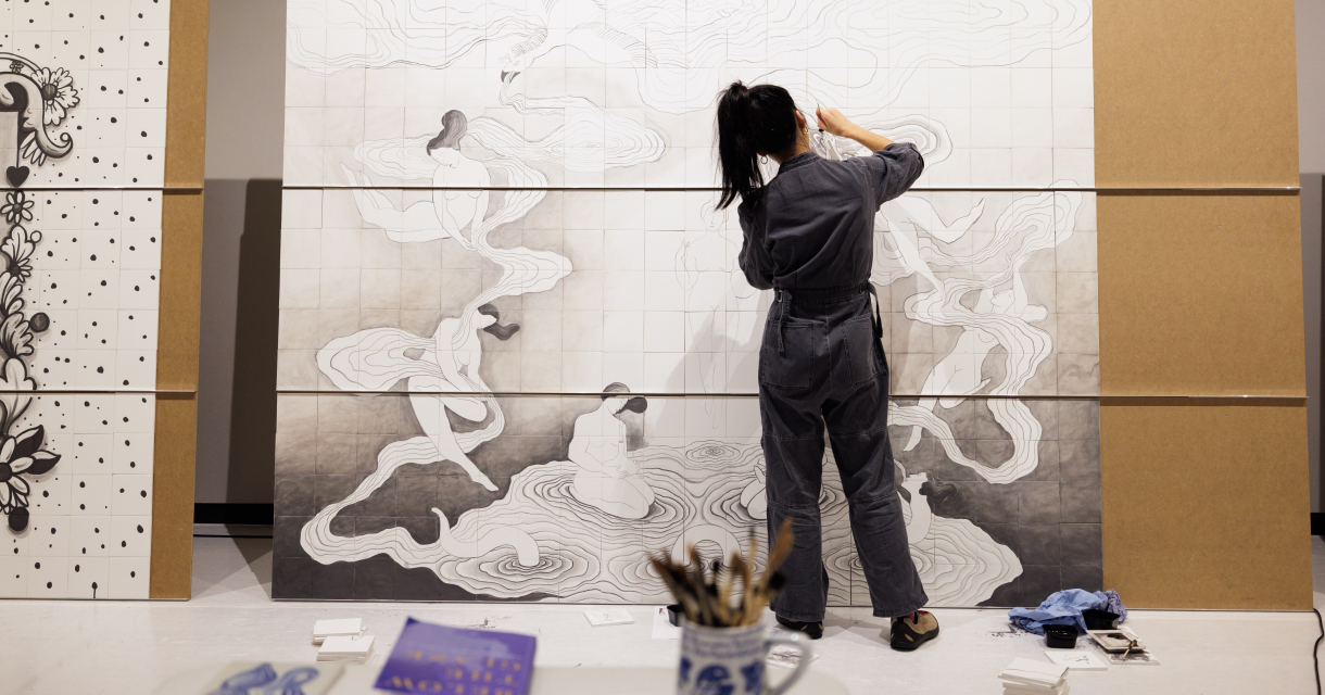 An artist is working on a large mural featuring female figures.