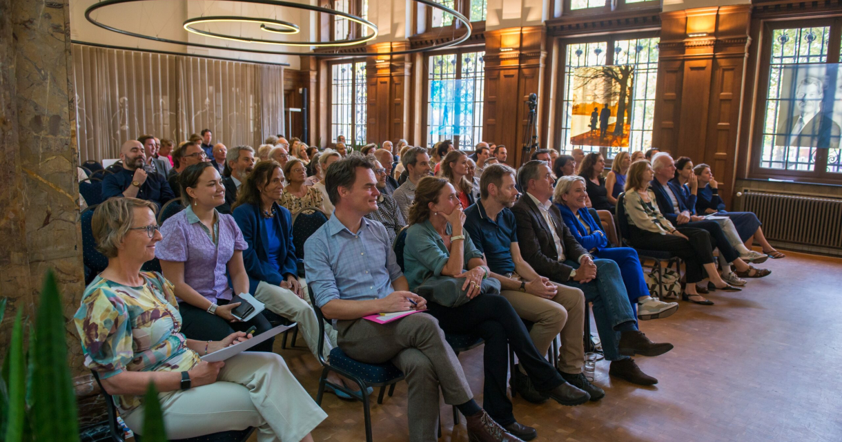 Een groep mensen zit in een evenementruimte, aandachtig luisterend.