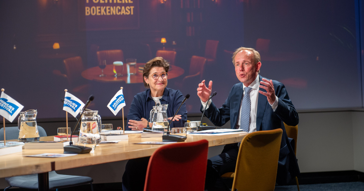 Twee sprekers discussiëren aan een tafel met boeken en glazen.