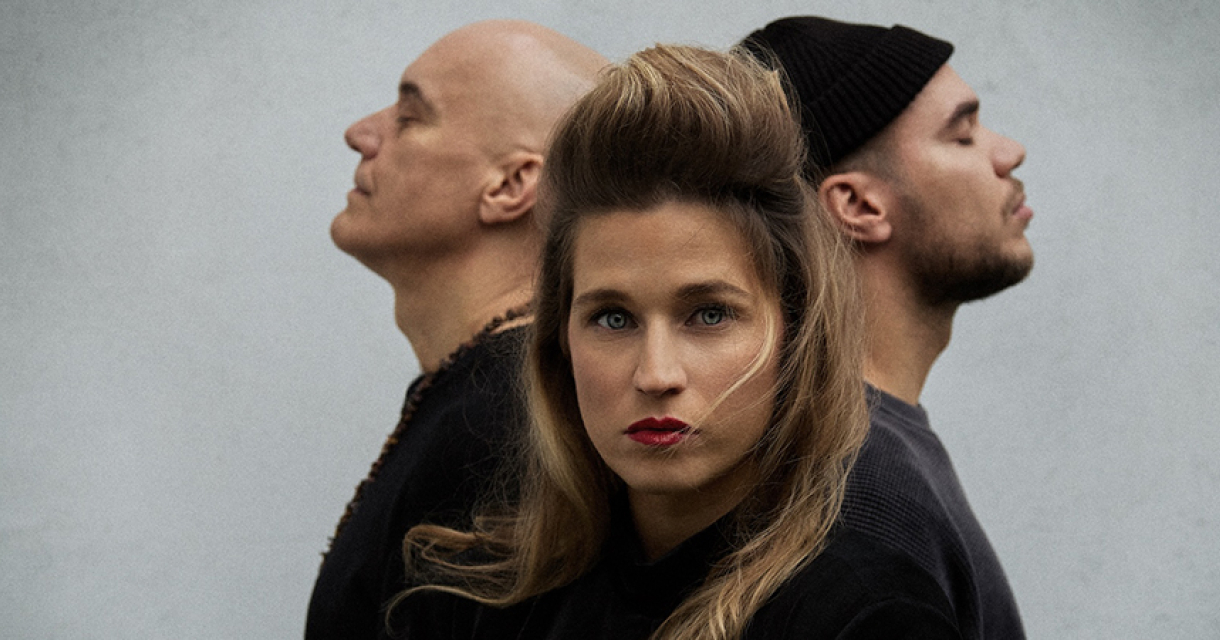 Drie personen met opvallende poses. Een vrouw met lange haren en rode lippen, met twee mannen in de achtergrond. De mannen zijn met hun rug naar de camera gekeerd.