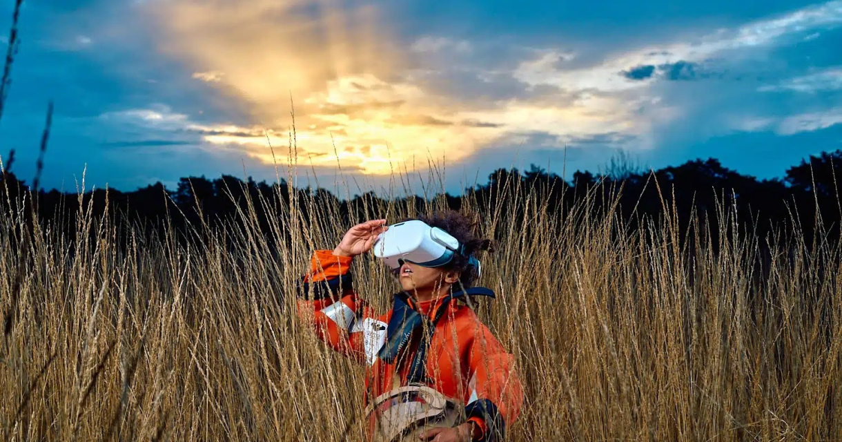 Installatie: ERGENS (8+). Een persoon met een VR-bril staat in hoog gras en kijkt naar de lucht tijdens zonsondergang.