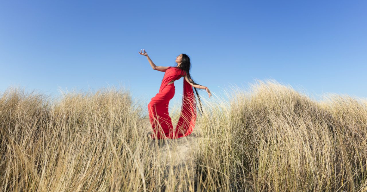 Een vrouw in een rode jurk poseert tussen hoge grassprieten onder een blauwe lucht.