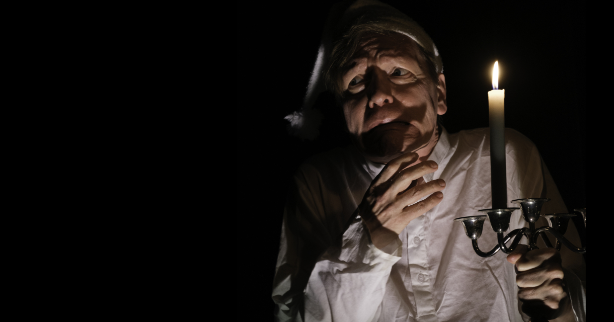 A man wearing a white nightcap and nightshirt anxiously holds a candlestick with a burning candle in the dark.