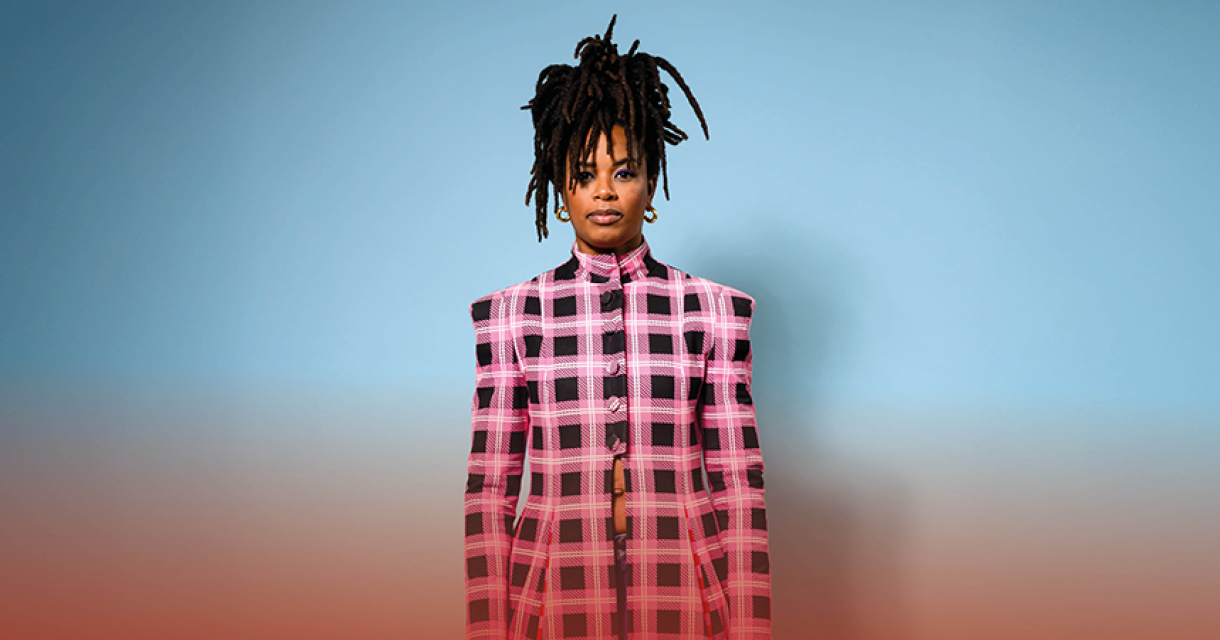 Shary-An in een kleurrijke outfit met dreadlocks, ze draagt een roze geruite jas voor een blauwe achtergrond.