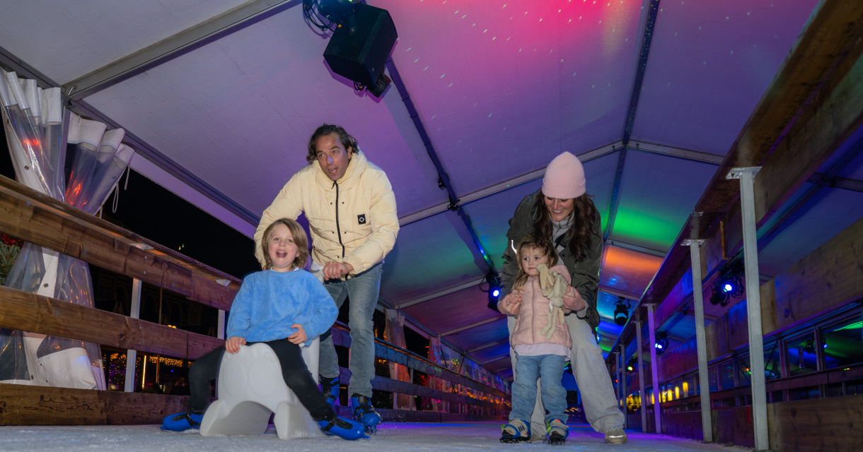 Een man helpt een kind met schaatsen, terwijl een vrouw een peuter vasthoudt op een ijsbaan.