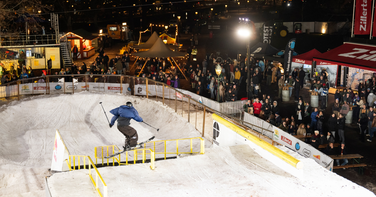 Un skieur saute par-dessus un obstacle dans un parc de neige temporaire avec un public en arrière-plan.
