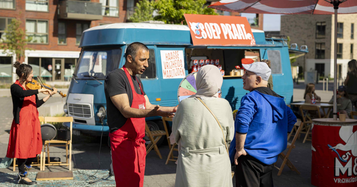 Een man in een schort praat met twee bezoekers op een markt.