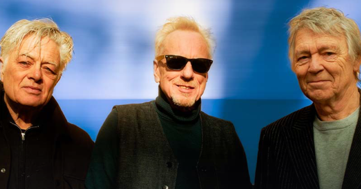 Drie mannen poseren met zonnebrillen tegen een blauwe achtergrond.