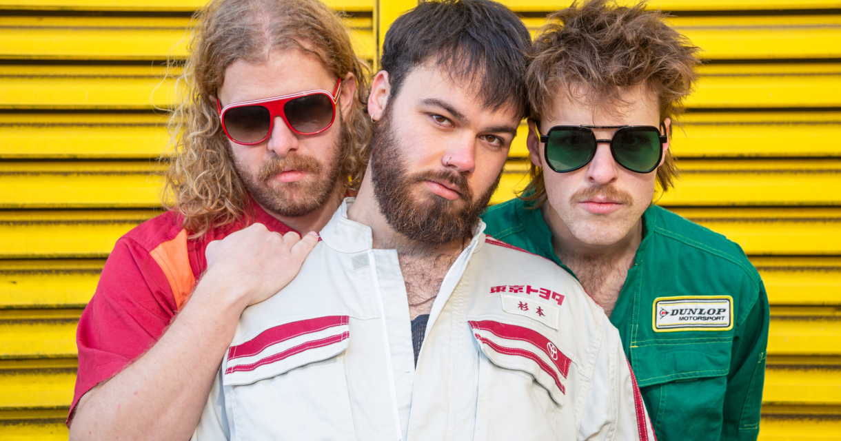 Drie mannen van de band TaxiTaxi poseren in kleurrijke kleding tegen een gele achtergrond.
