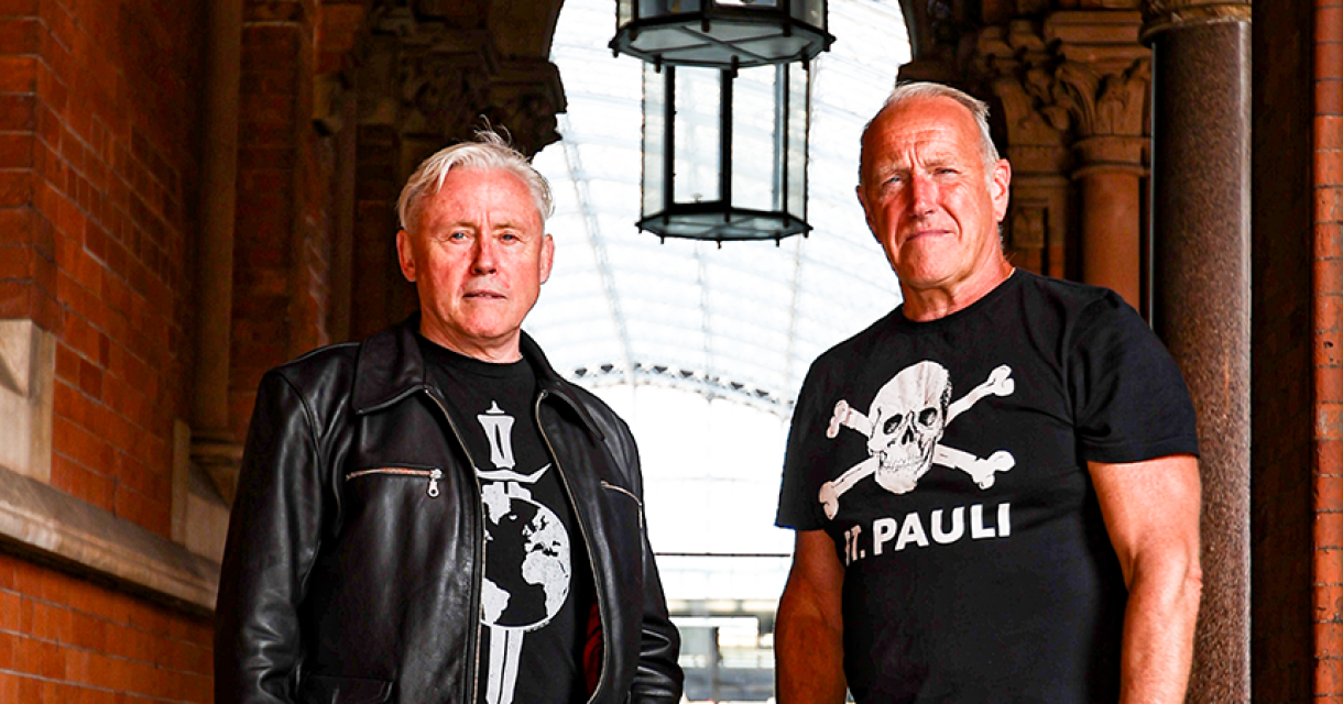 Kirk Brandon en Stan Stammers van Theatre of Hate staan samen in een gebouw met bakstenen muren.