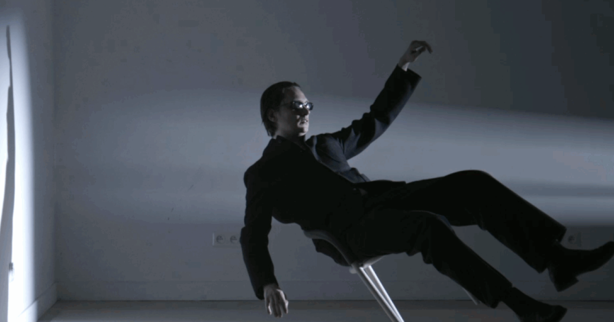 Thomas Azier in een zwart pak leunt achterover op een stoel en kijkt naar de schaduw op de muur.