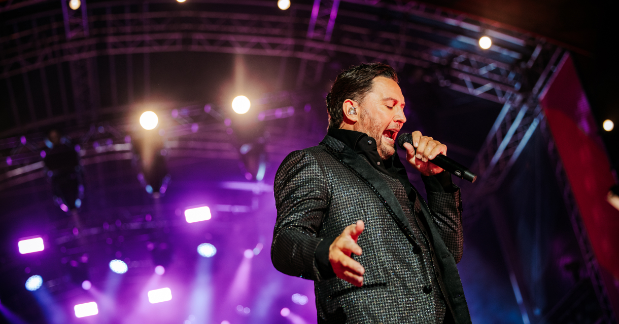 Een zanger in een glanzend kostuum zingt intens op een podium met gekleurde lichten.