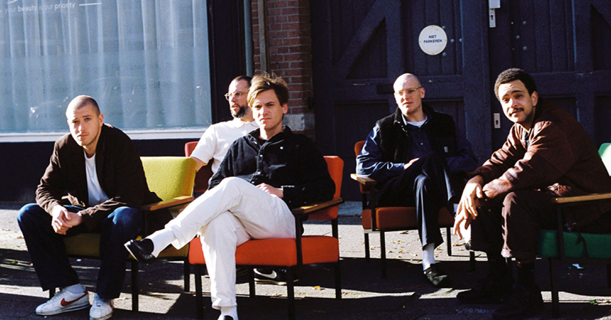 Vijf mannen zitten op kleurrijke stoelen voor een gebouw.