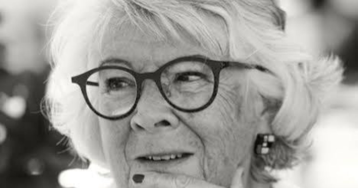 Vrouw met grijs haar en bril, glimlachend terwijl ze nadenkt.
