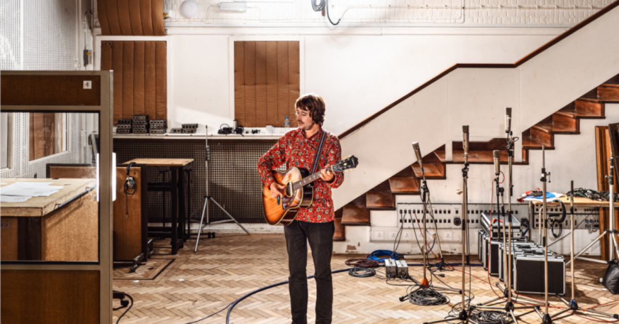 Een man speelt gitaar in een muziekstudio met microfoons en apparatuur.