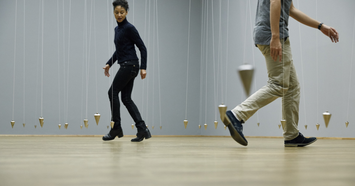 Two people walking among hanging pendulums in a gallery.