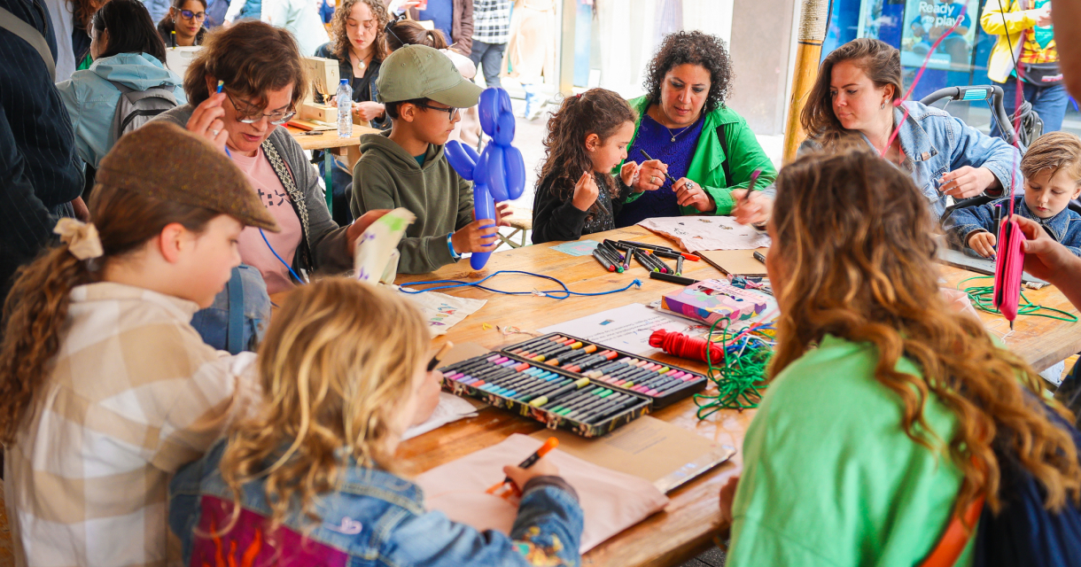 Groep kinderen aan een grote tafel vol knutselspullen die een workshop doen onder begeleiding van twee volwassen vrouwen.