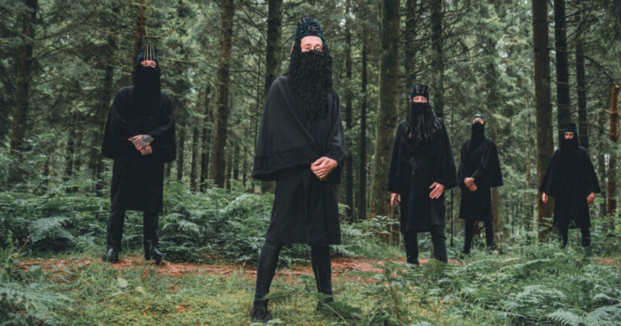 Vijf personen van band Wyatt E in zwarte outfits staan in een bosomgeving.