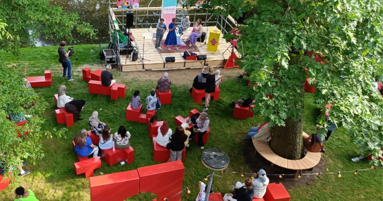 Een buitenoptreden met een publiek op rode stoelen.