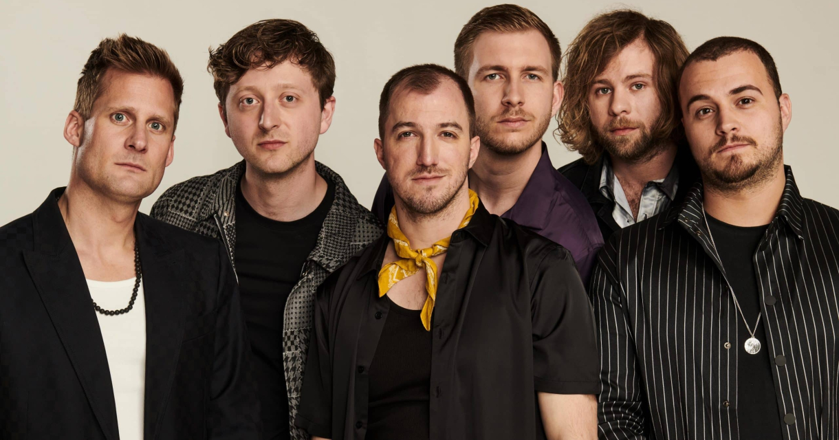 Zes mannen poseren samen met verschillende kleurrijke outfits.