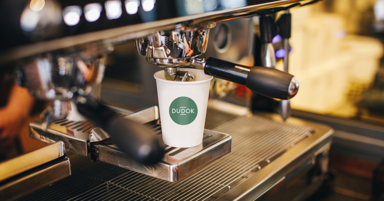 Kartonnen beker met logo van Dudok onder een professioneel koffieapparaat. 