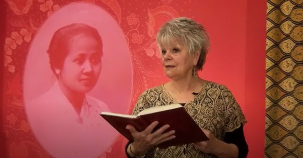 Anita Poolman draagt voor met een boek in haar hand, staand voor een portret van Kartini; dochter van de regent van Japara