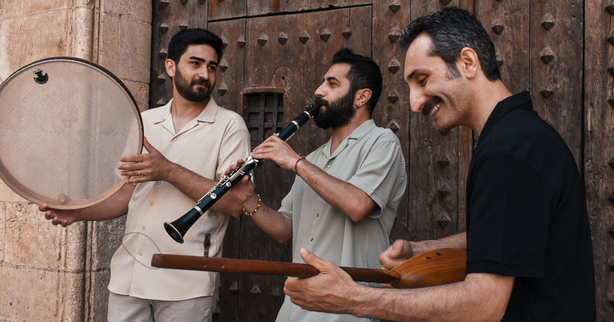 Drie mannen spelen traditionele instrumenten buiten voor houten deur: trommel, klarinet en snaarinstrument