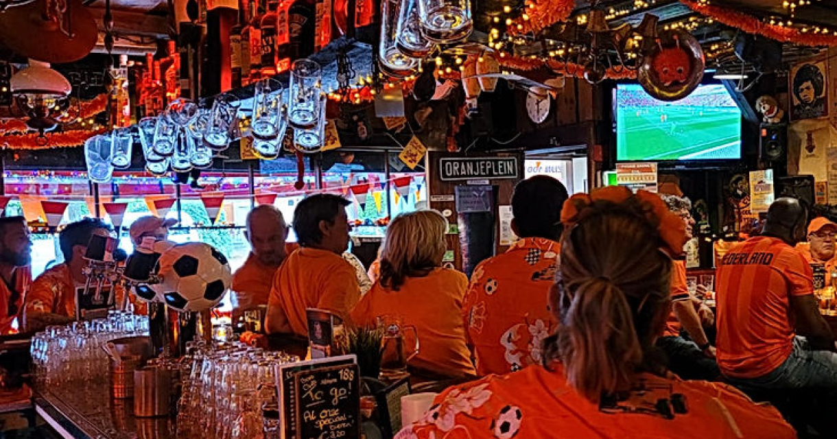 Mensen in oranje shirts kijken naar een wedstrijd in een bar, met versiering en een tv.