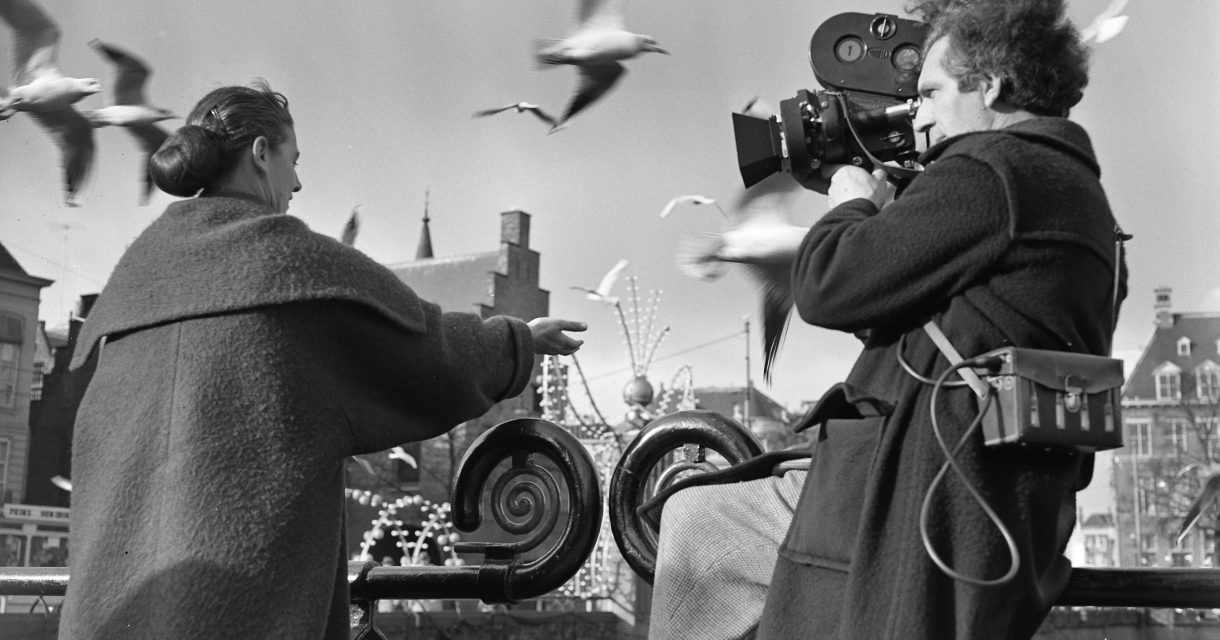 Historische zwart wit foto van een man die met een 8 mm camera een vrouw filmt die meeuwen voert