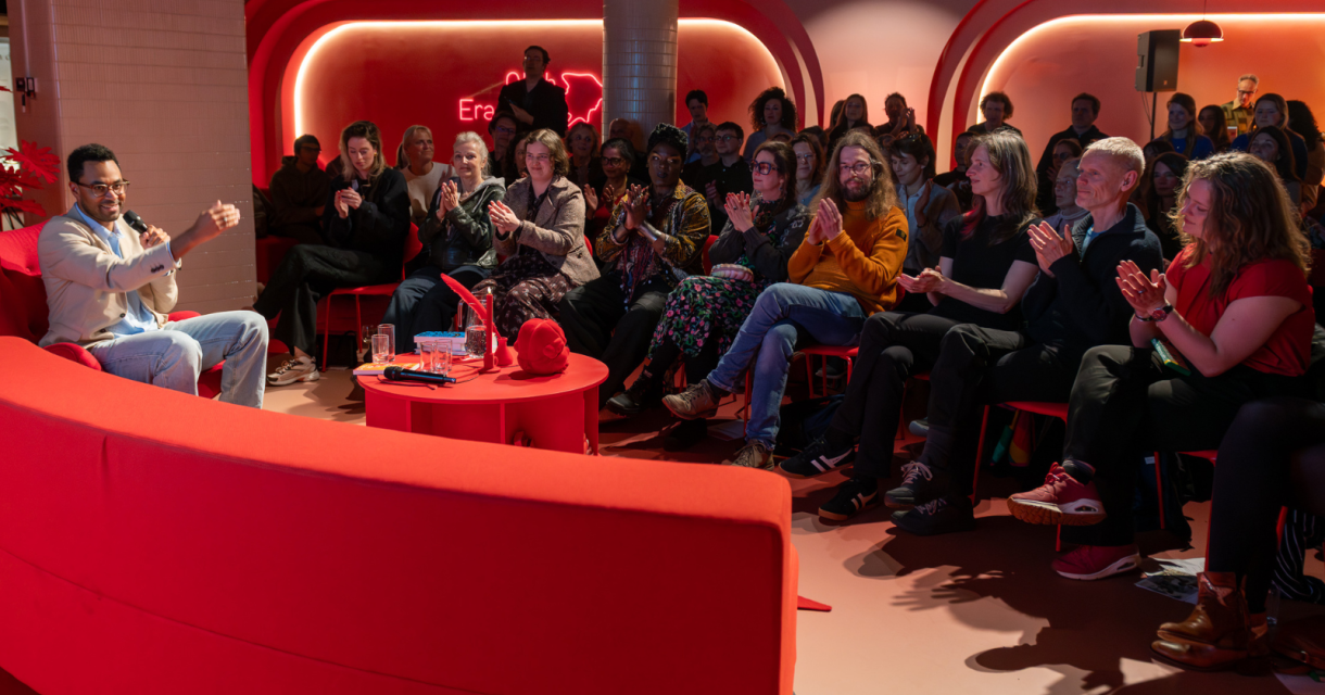 Een spreker praat in een felrode omgeving met een applaudisserend publiek.