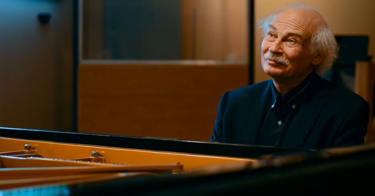 Een man met grijs haar zit met een glimlach achter een piano.