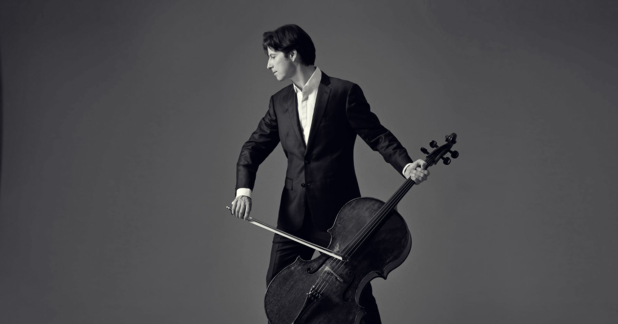 Een cellist in een zwart pak, naast zijn cellokoffer, met een strijkstok in hand.