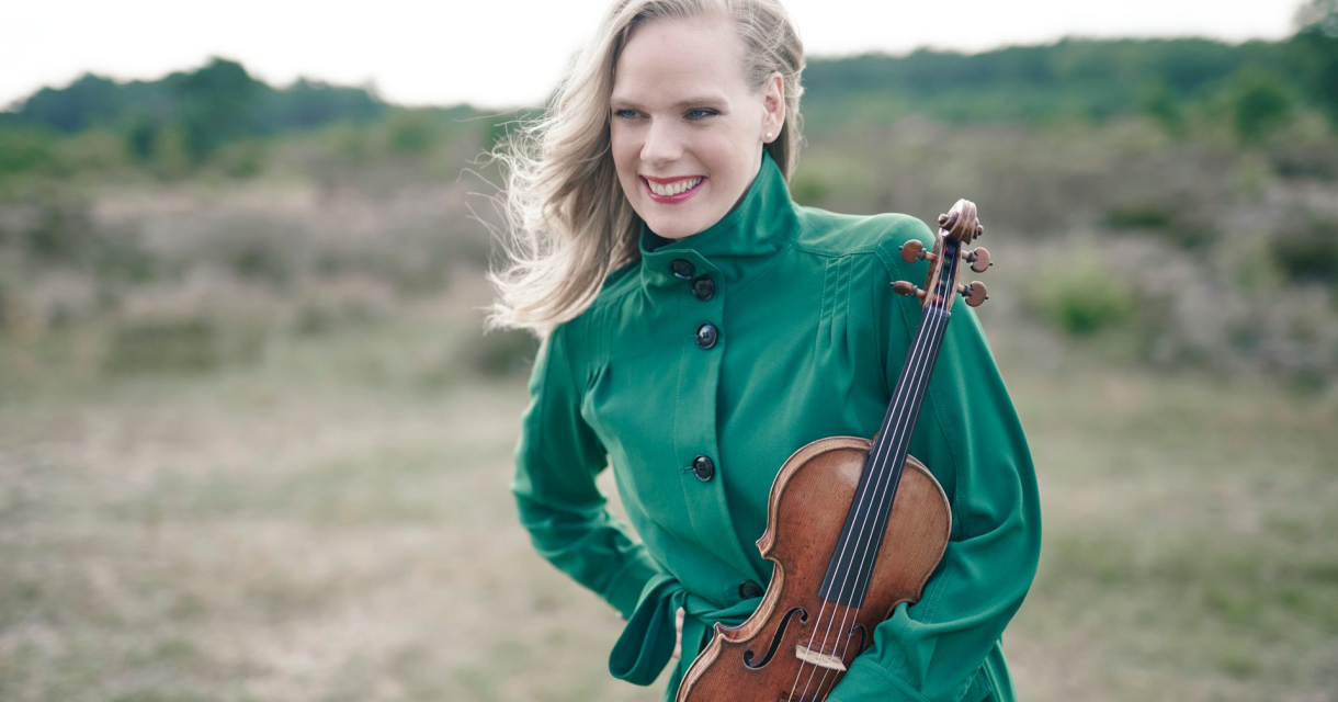 Een vrouw met een groene jas houdt een viool vast en glimlacht.