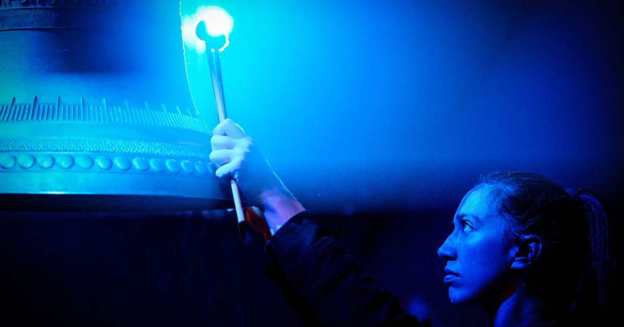 Een vrouw raakt een grote bel aan met een hamer in een blauwe omgeving.