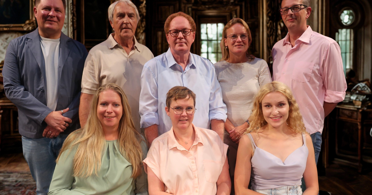 Een groep van acht mensen poseert voor een verlichte, antieke achtergrond.