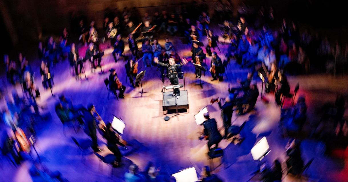 Een dirigent leidt een orkest in een zaal met blauwe lichten.