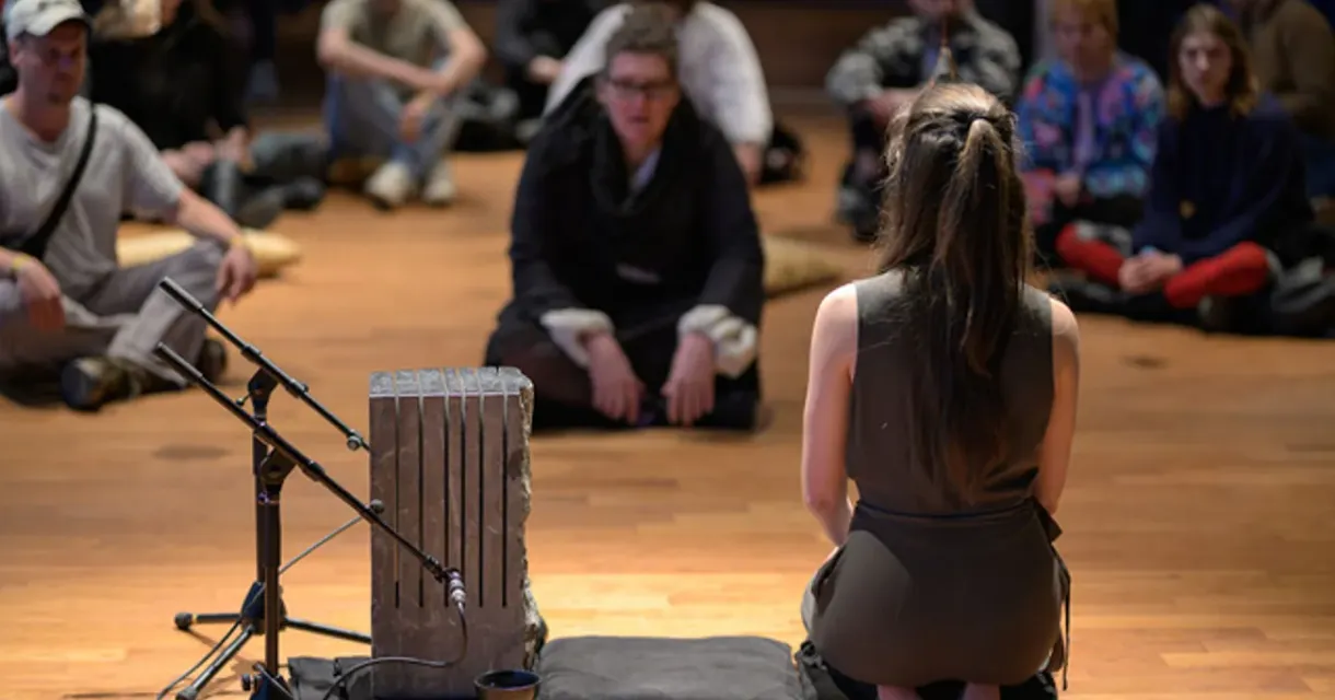 A woman performs for a group of people, with a musical instrument in front of her.