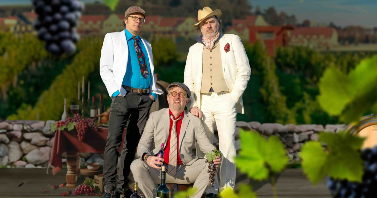 Drie mannen poseren naast een tafel met wijn en druiven.